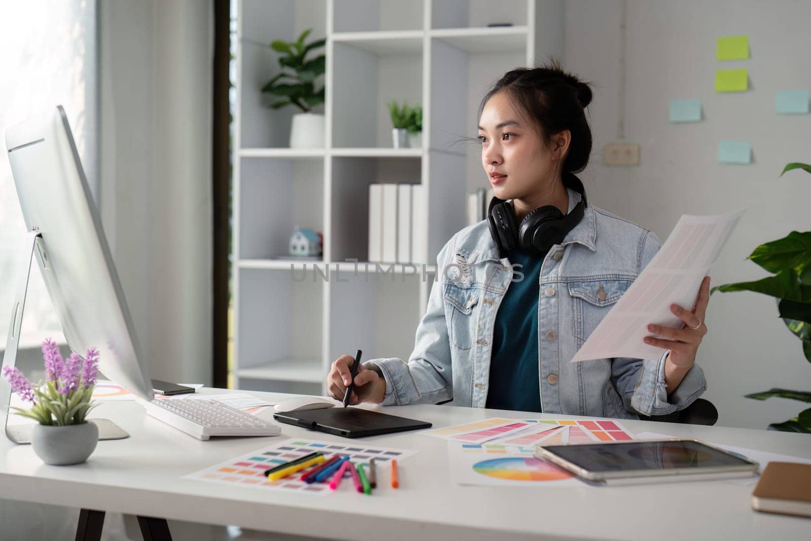 Asian woman freelance graphic designer working with color swatch samples and computer at desk in home office, young lady choosing color gamma for new design project.