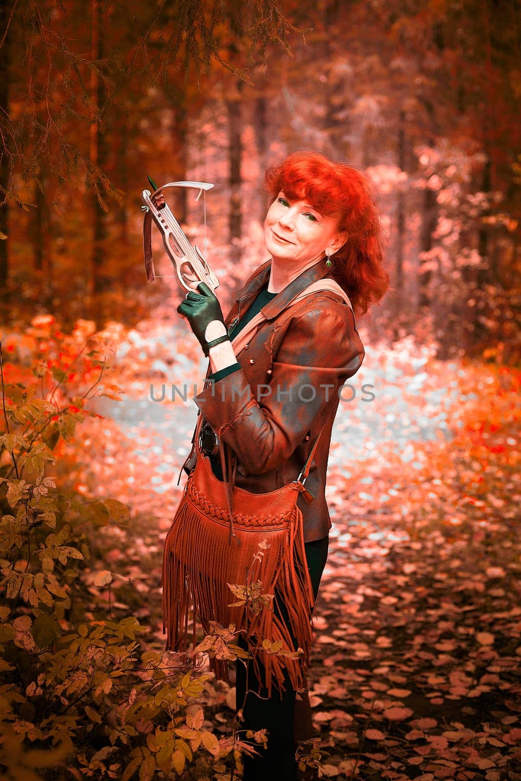 Mature model portraying a royal huntress with red curve hair is hunting with a crossbow in the in vibrant autumn forest in thematic photo shoot