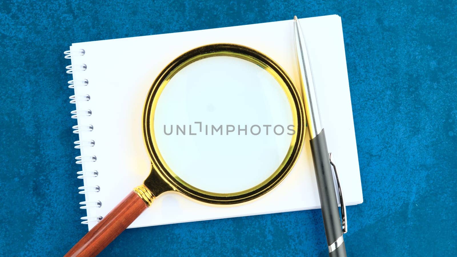 A photo on a blue background with a copy space with a notepad, magnifying glass and pen in the foreground. Concept photo by Ihar