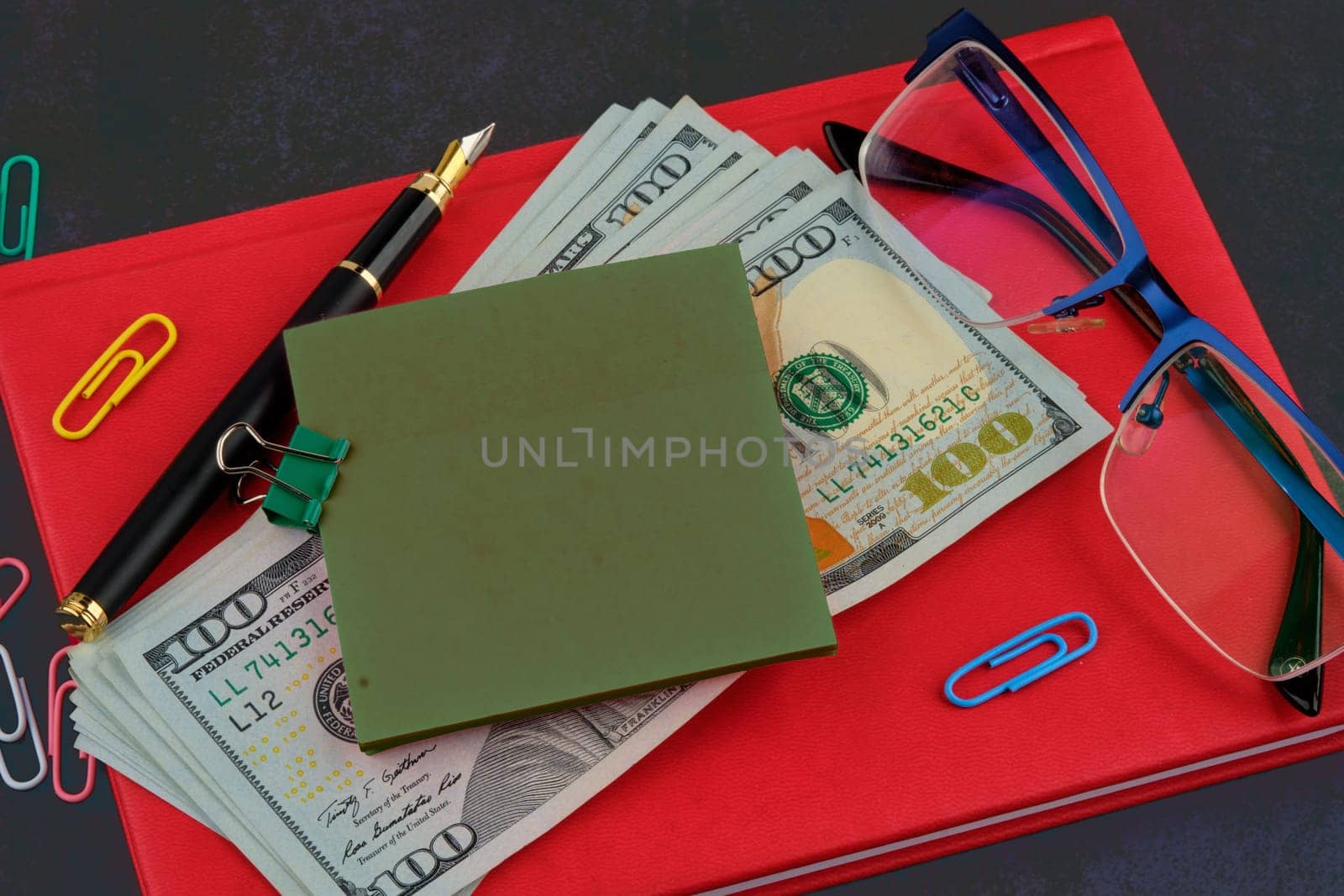 A sticker for writing attached to dollars on a red business notebook, glasses next to it, a pen on a dark background by Ihar