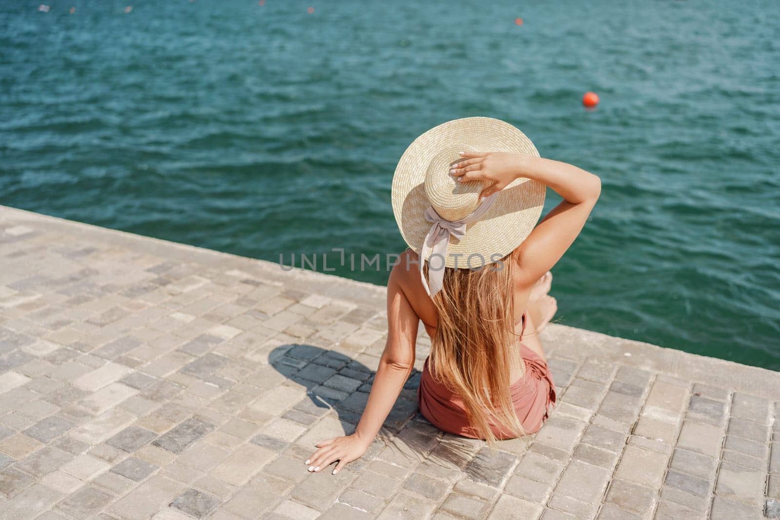 A woman in a swimsuit sits with her back holding a hat, looks at the ocean, sunny day, relaxes. by Matiunina