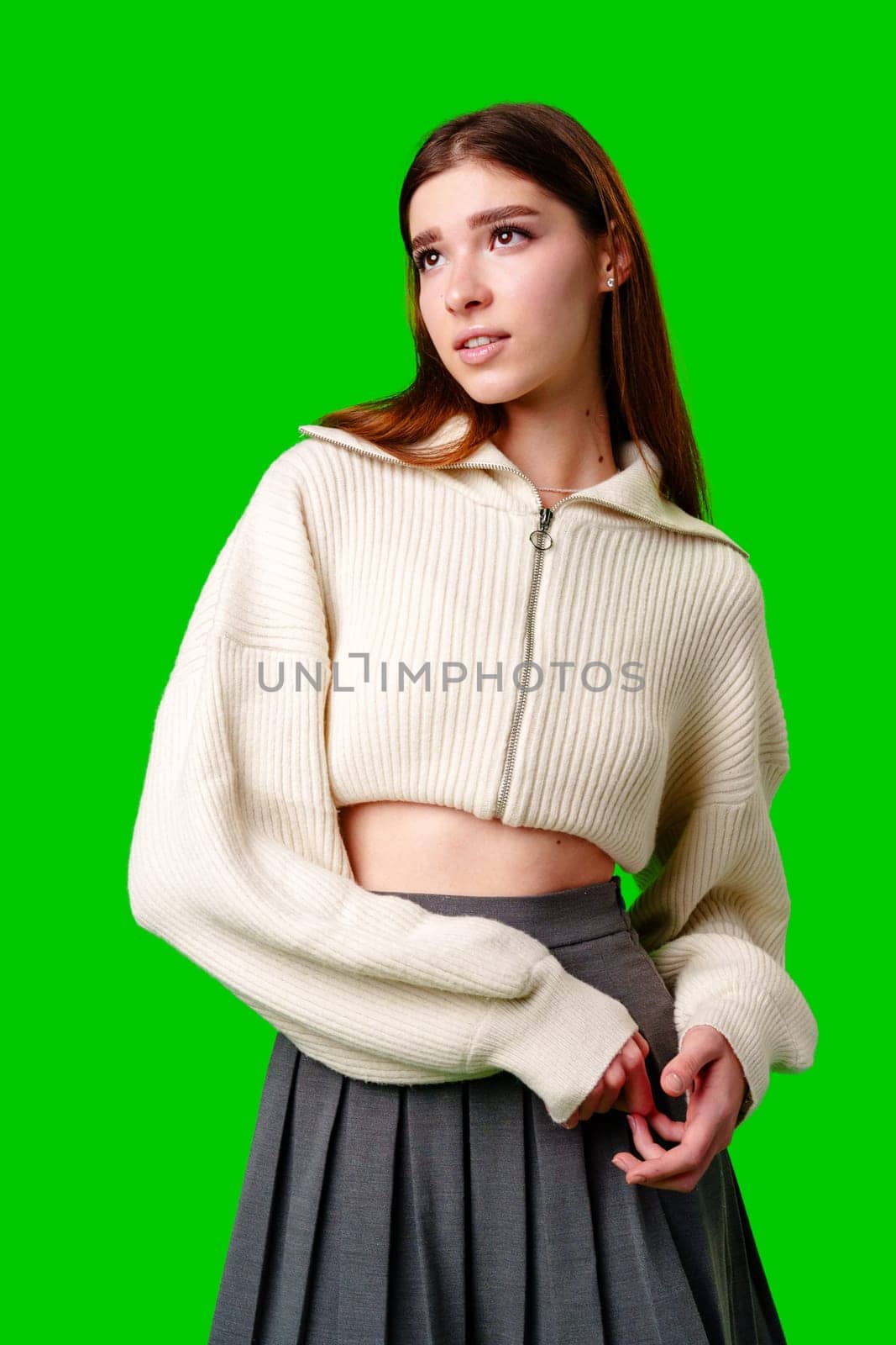 A young woman stands against a vibrant green backdrop, her gaze directed off-camera with a thoughtful expression. She wears a casual, cropped white zip-up sweater paired with a dark pleated skirt, and her long brown hair hangs straight over her shoulders.