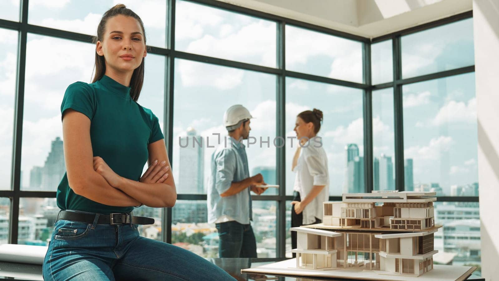 Young beautiful interior designer crossing arms while engineer team talking about house design. Skilled businesswoman smiling at camera with confident while sitting near house model. Tracery