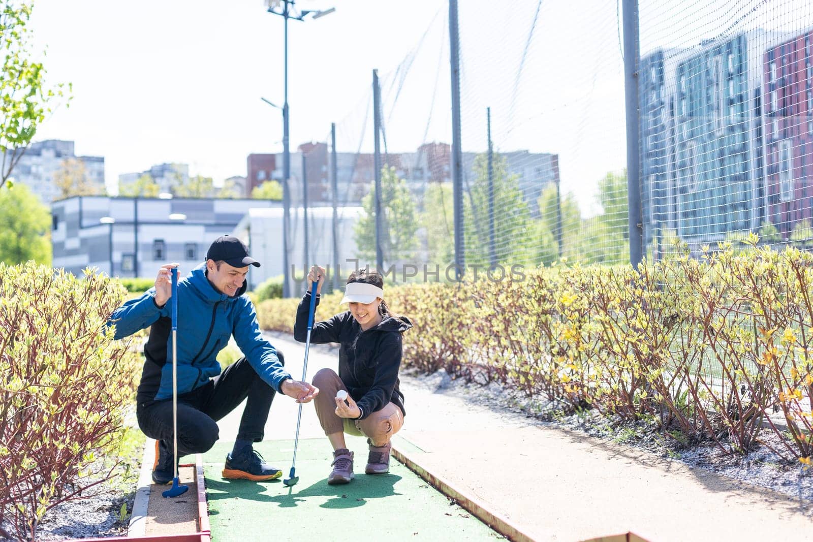 The girl is preparing to hit the ball with a golf club and looks at the man who is squatting next to her with the golf club in his hands. High quality photo