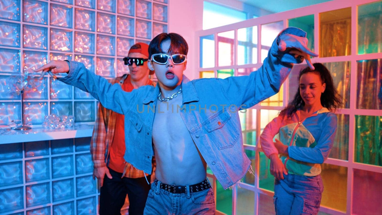 Asian man moving his arm while dancing with diverse team in neon light at night club. Happy dancer with stylish cloth moving along with multicultural hipster in lively mood at modern club. Regalement.