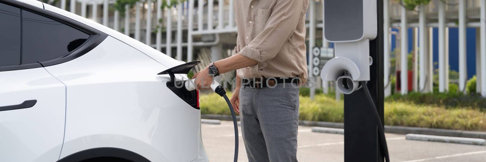 Young man recharge electric car's battery from charging station in city commercial parking lot. Rechargeable EV car for sustainable environmental friendly urban travel. Panorama Expedient