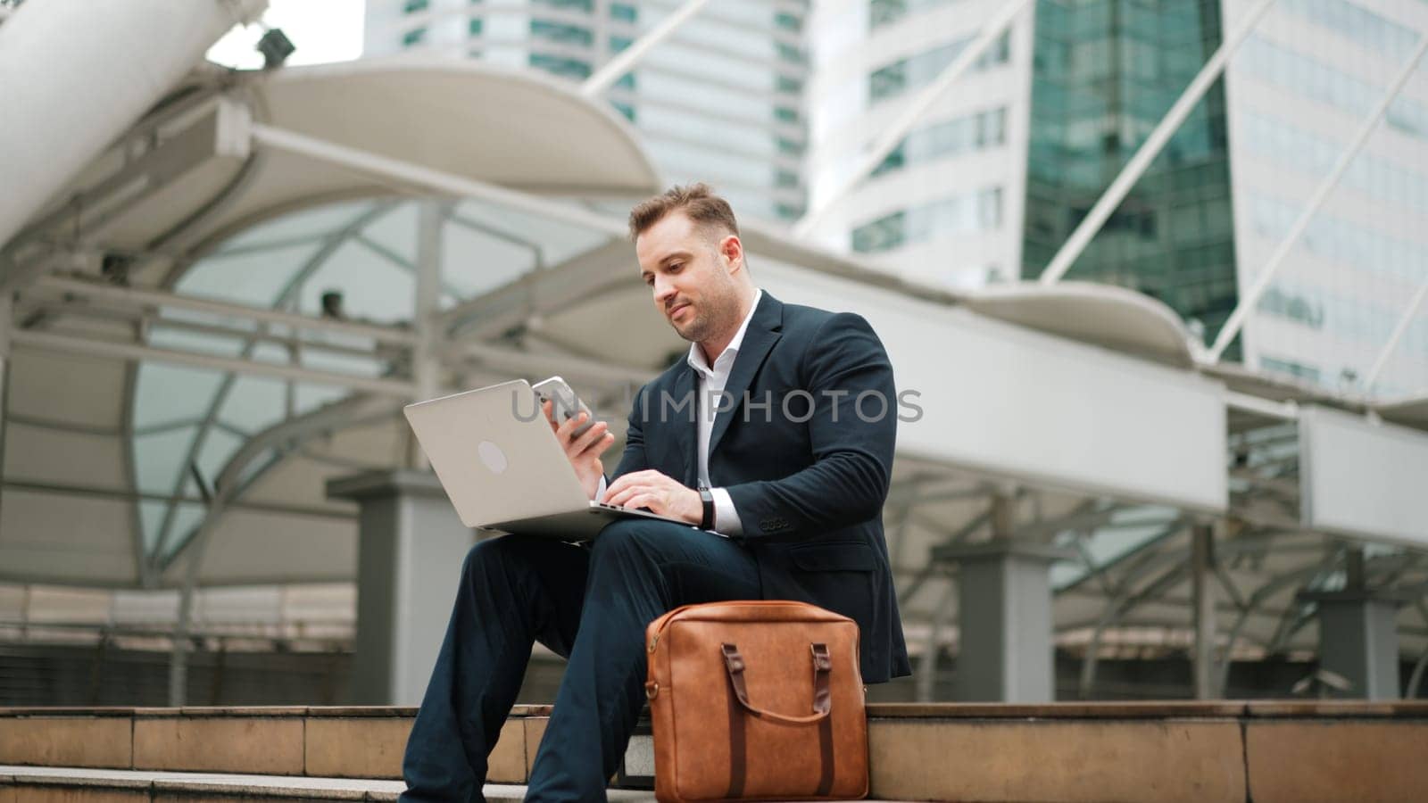 Smart caucasian business man sitting at stairs while using laptop to type to project manager and calling phone to present and plan marketing strategy surrounded by urban city view. Lifestyle. Urbane.