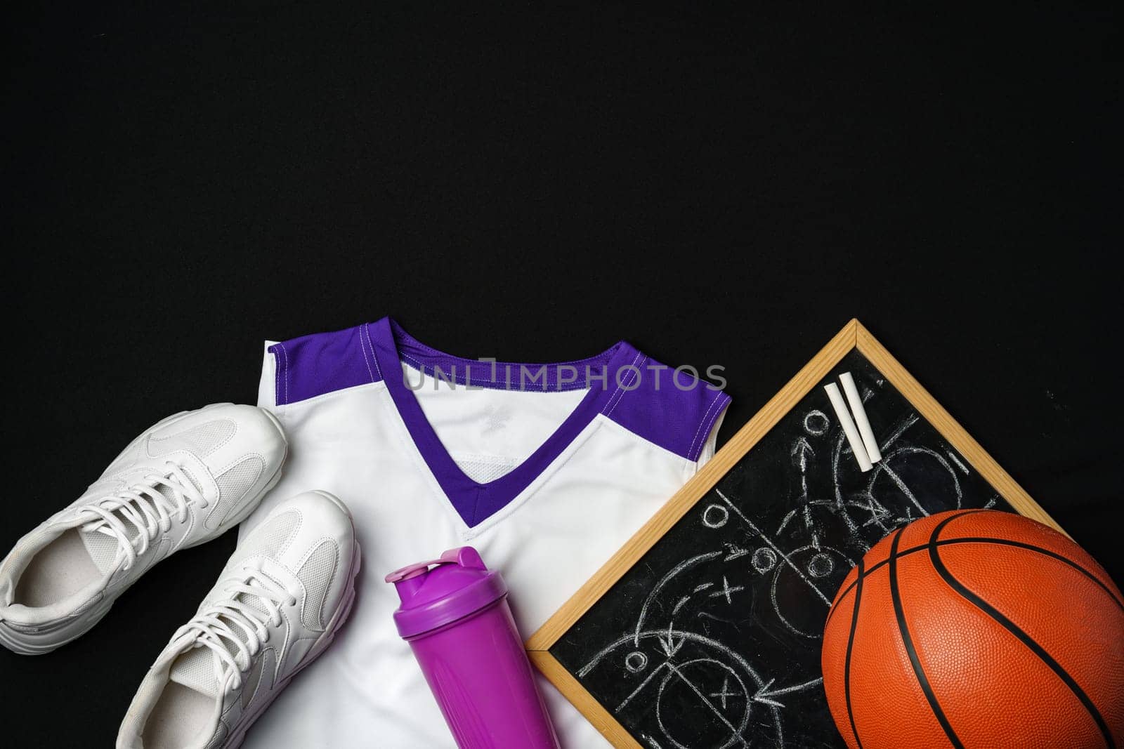A collection of basketball essentials is neatly arranged against a stark black backdrop. A pair of white sneakers with purple accents lies next to a vibrant orange basketball. On the right, a clipboard with a basketball court diagram and a strategy sketched in white chalk can be seen, while a white and purple basketball jersey drapes gracefully at the edge. Completing the set, a purple sports water bottle caps off the ensemble, suggesting preparation for an upcoming game or practice session.