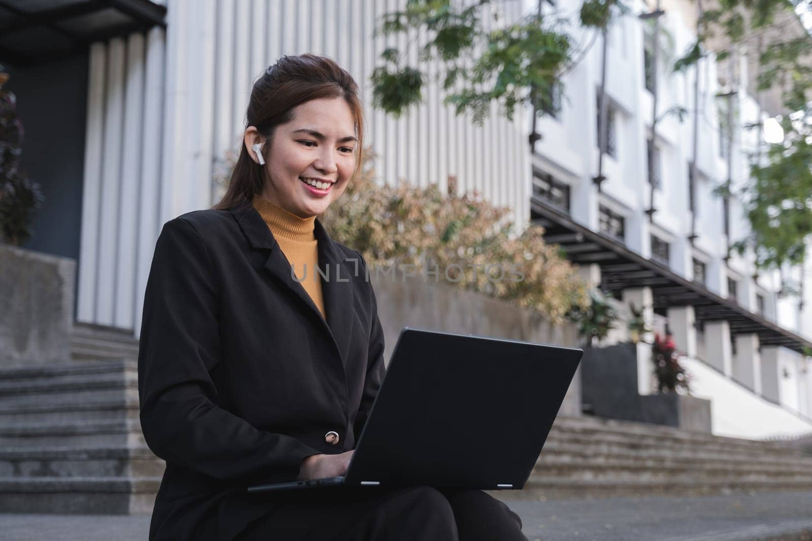 A woman in a black suit is sitting on a step with a laptop in front of her. She is smiling and she is enjoying her work