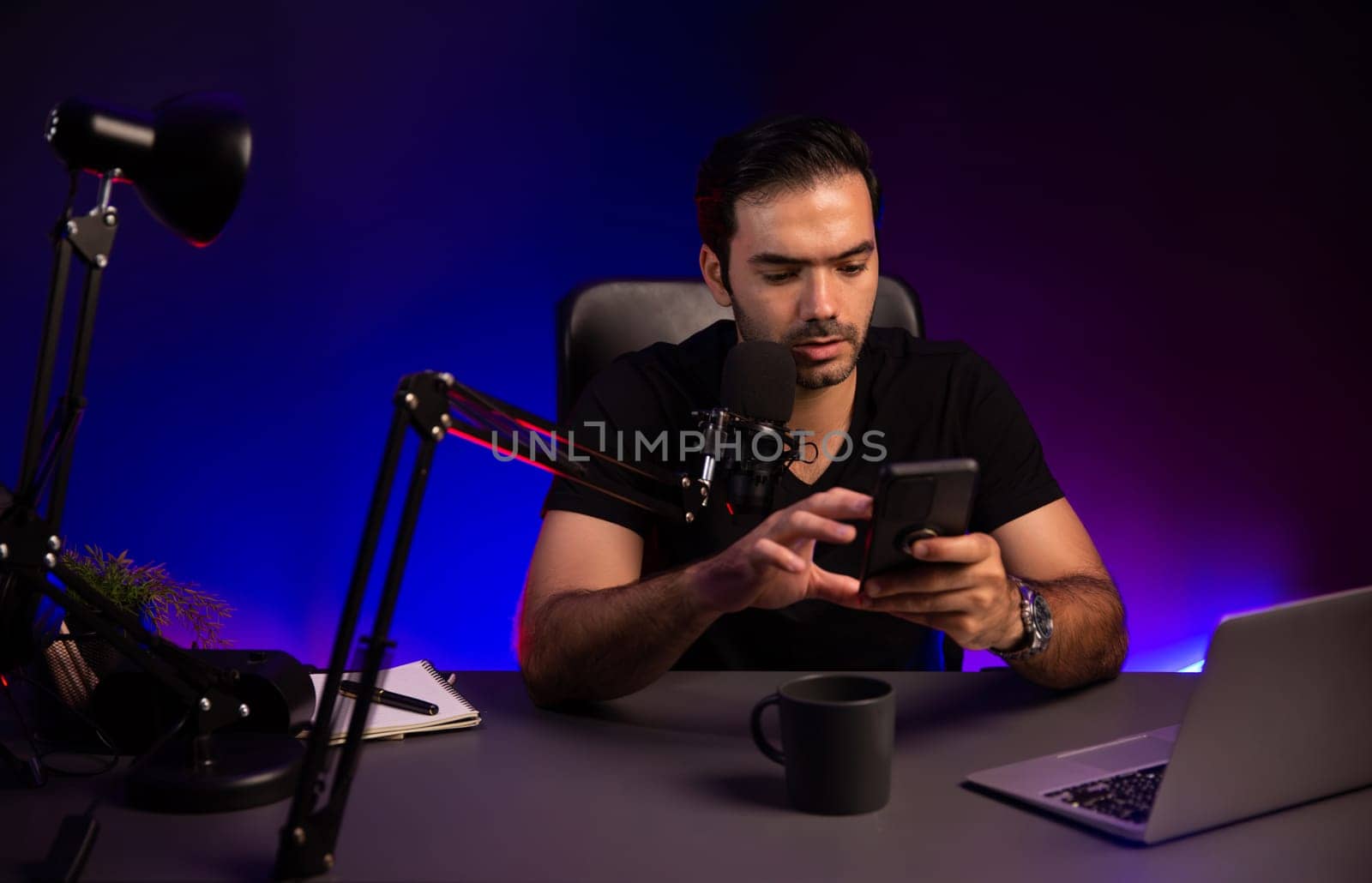 Broadcaster talking to listener on live streaming online, checking message from smartphone on social media in host channel by creative positive thinking concept at purple neon light studio. Surmise.