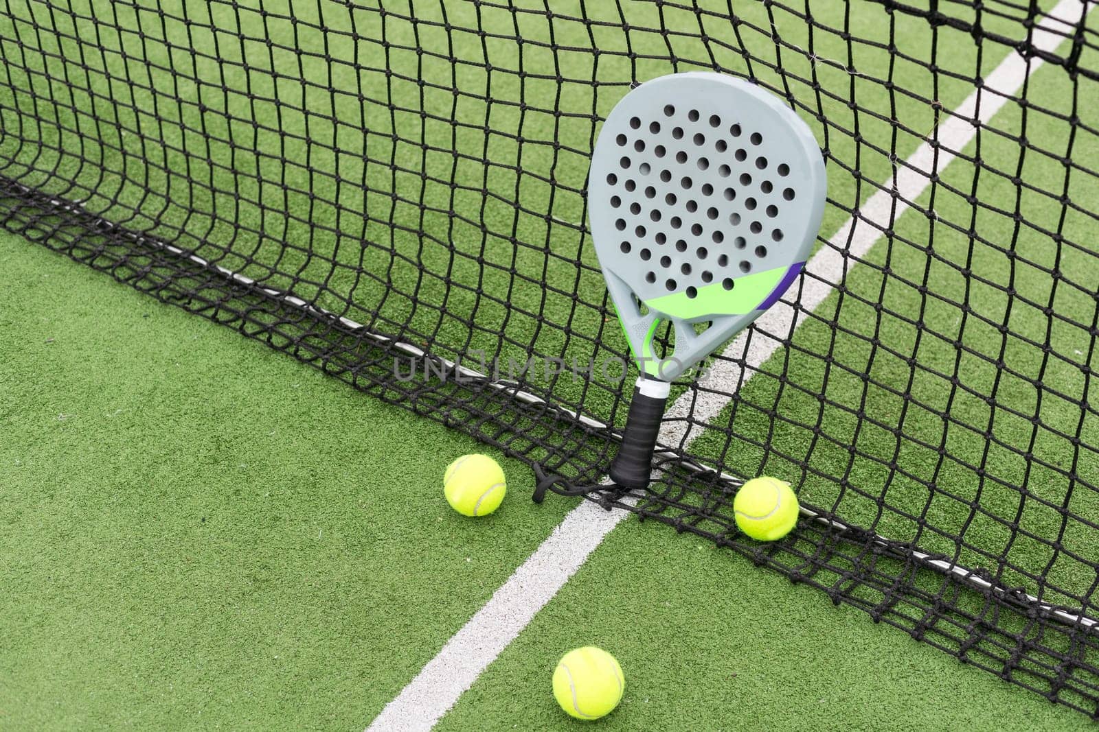 Yellow balls on grass turf near padel tennis racket behind net in green court outdoors with natural lighting. Paddle is a racquet game. Professional sport concept with copy space. High quality photo