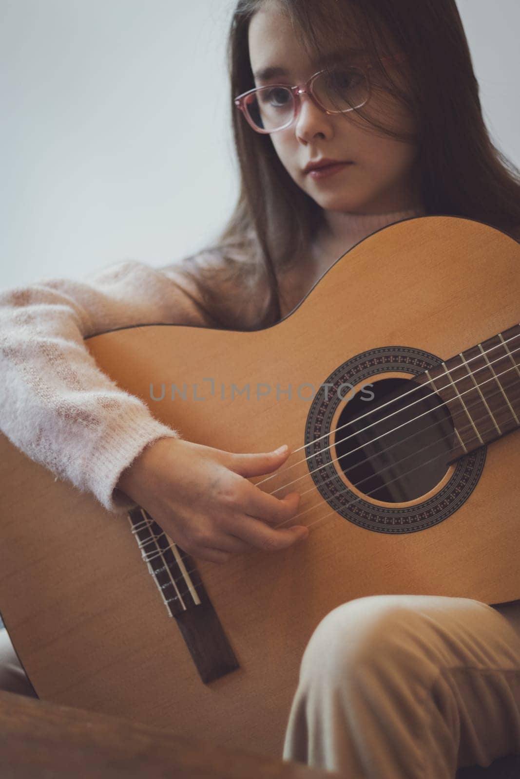 A beautiful little caucasian brunette woman with flowing long hair in glasses holds a guitar and plucks the strings with her fingers, looking at the notes on the table, sitting on the sofa in the room, close-up side view. Music education concept.