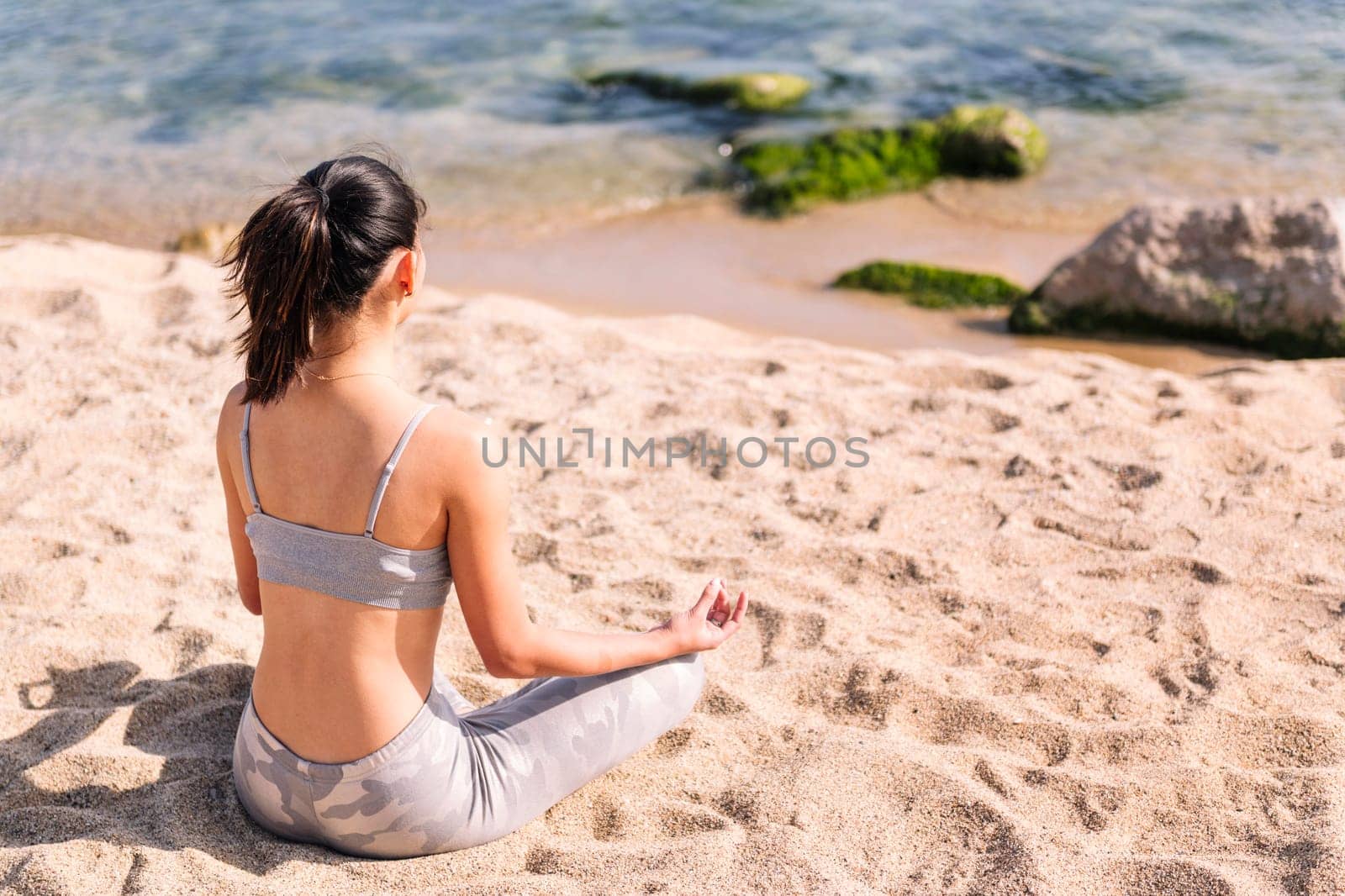 unrecognizable young woman in sportswear doing meditation at beach sitting with legs crossed, concept of mental relaxation and healthy lifestyle