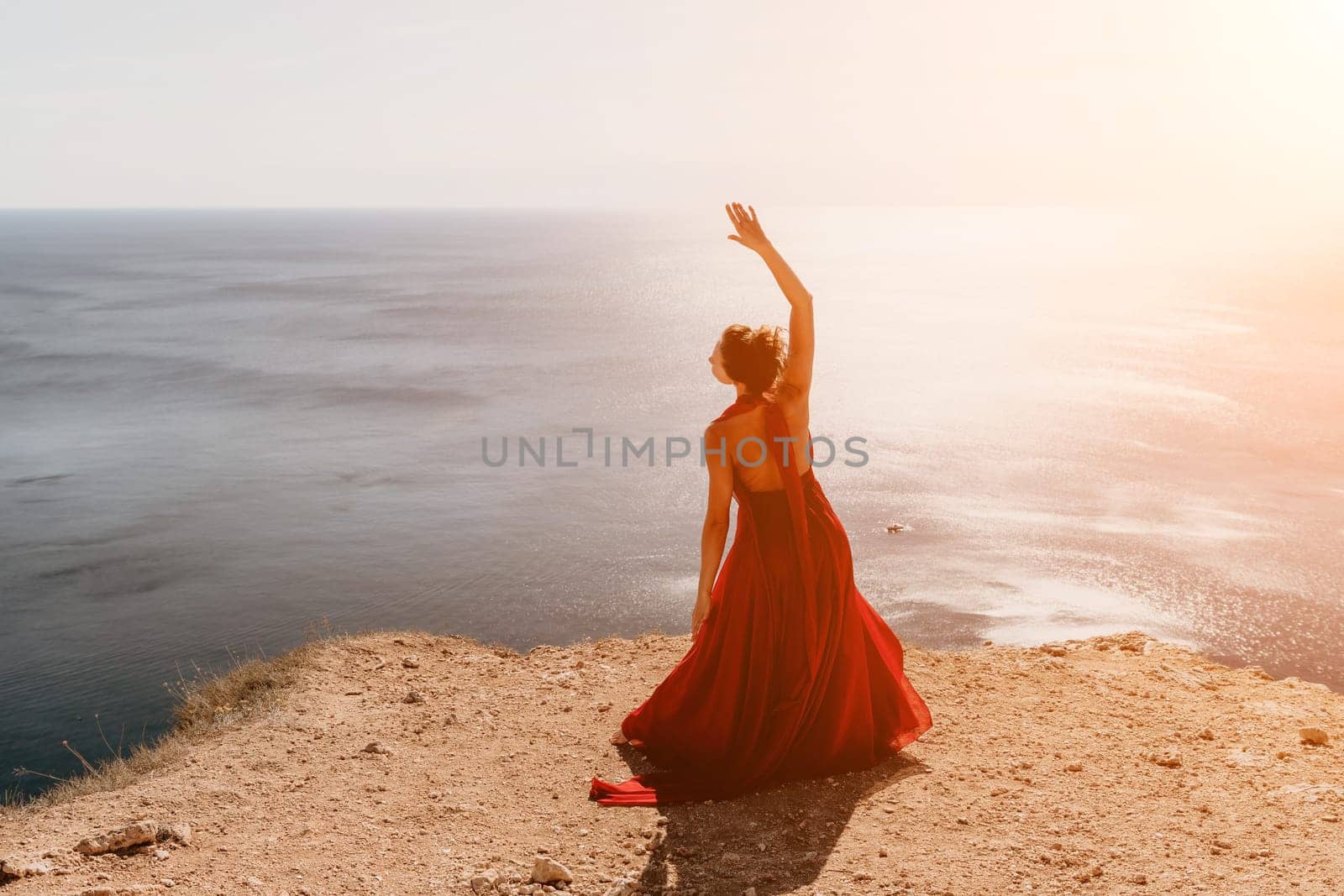 Side view a Young beautiful sensual woman in a red long dress posing on a rock high above the sea during sunrise. Girl on the nature on blue sky background. Fashion photo.