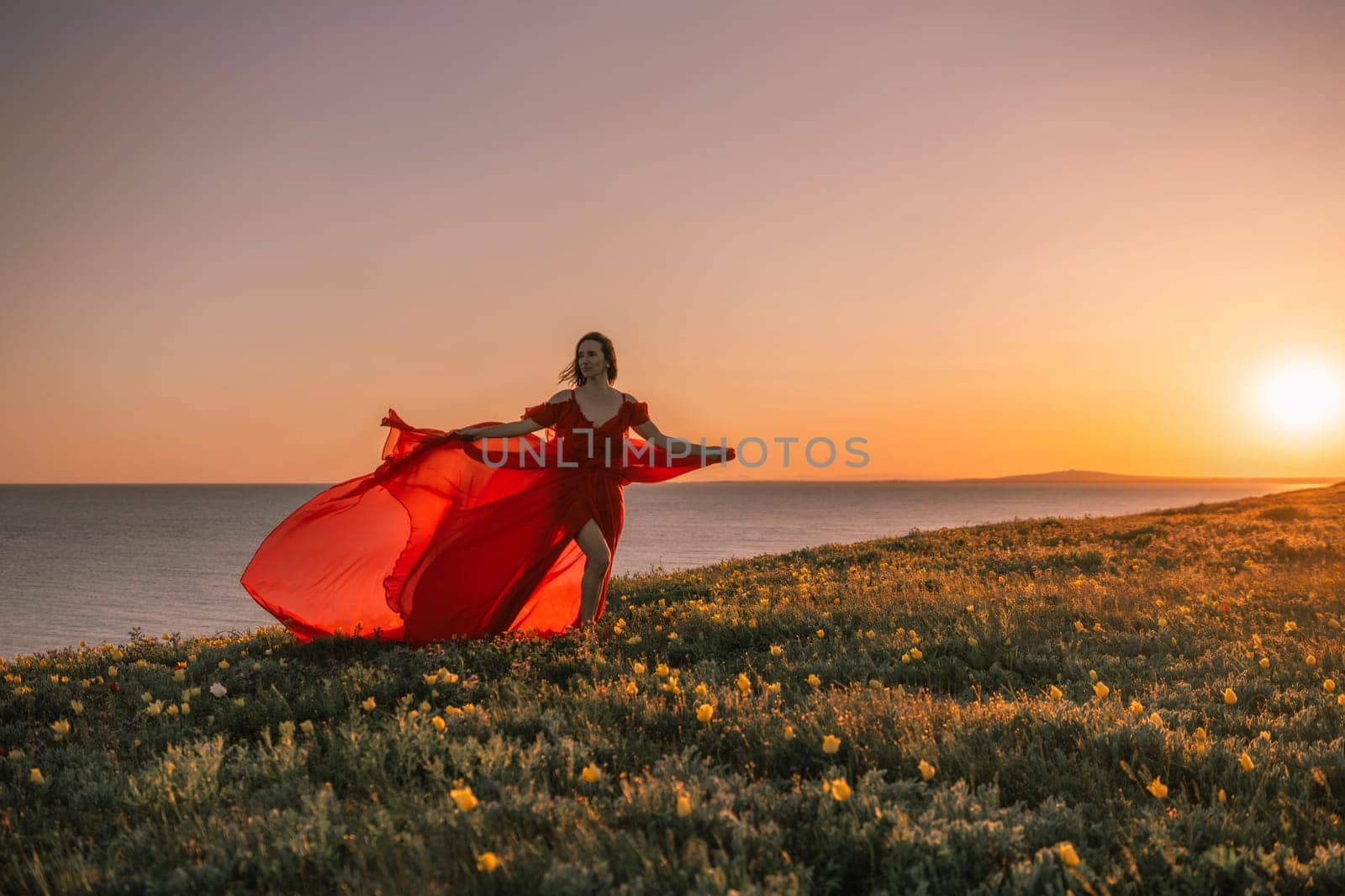 woman red dress is standing on a grassy hill overlooking the ocean. The sky is a beautiful mix of orange and pink hues, creating a serene and romantic atmosphere. by Matiunina