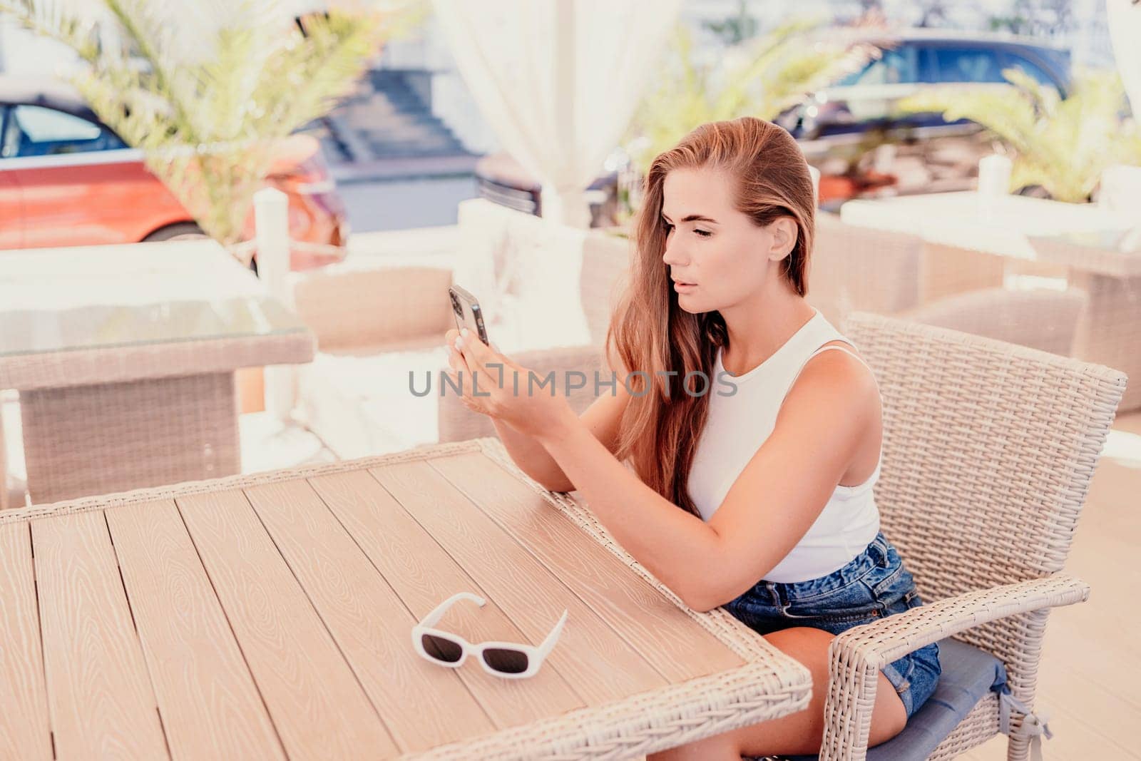Woman sits in outdoor cafe at wooden table, holds smartphone. Bright sunny day provides natural lighting. Cafe offers relaxed atmosphere for customers to enjoy refreshments. by Matiunina