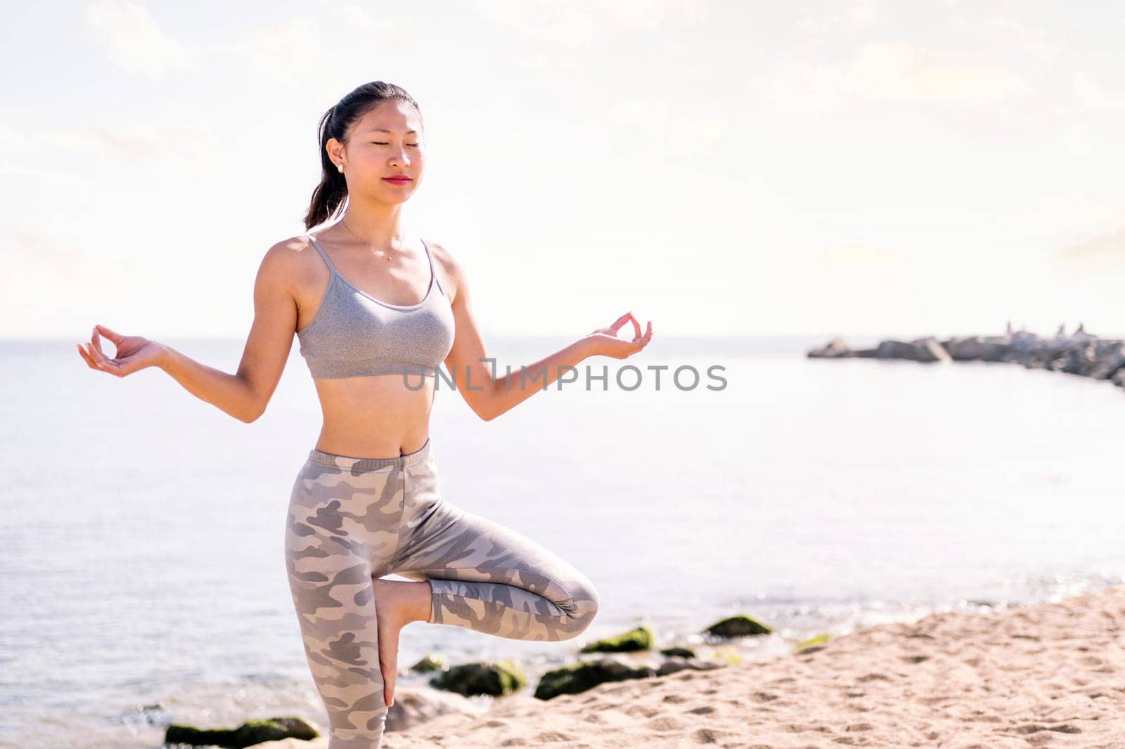 young asian woman in sportswear doing yoga position at beach, concept of mental relaxation and healthy lifestyle