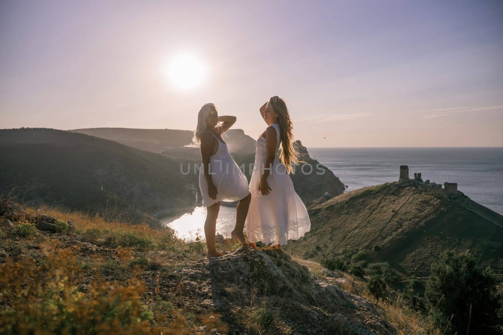 Two young girls are standing on a hillside, one of them wearing a white dress. The sun is shining brightly, creating a warm and inviting atmosphere. The girls seem to be enjoying their time together. by Matiunina