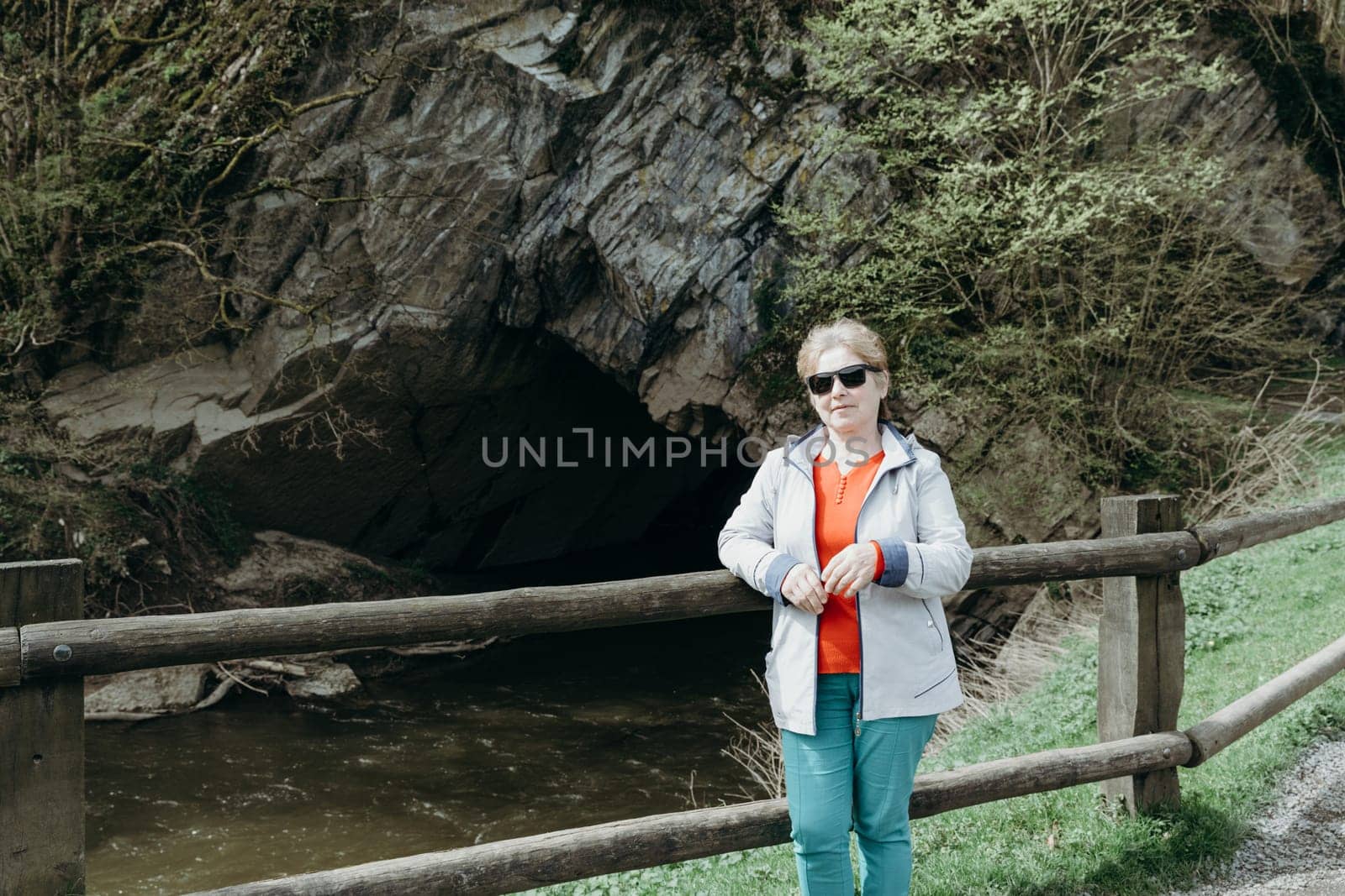 Portrait of a beautiful Caucasian elderly woman in sunglasses standing against the backdrop of a mountain cave with a river flowing into it on a sunny spring day in a nature reserve, close-up side view.