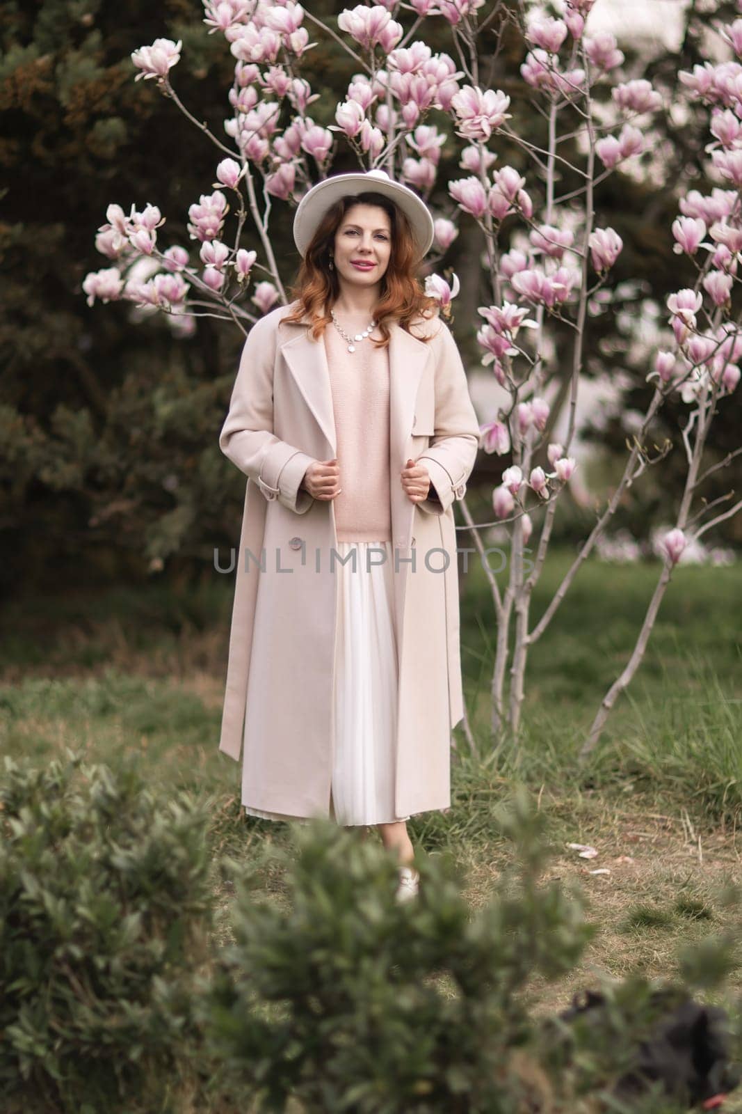 Woman magnolia flowers, surrounded by blossoming trees., hair down, white hat, wearing a light coat. Captured during spring, showcasing natural beauty and seasonal change. by Matiunina