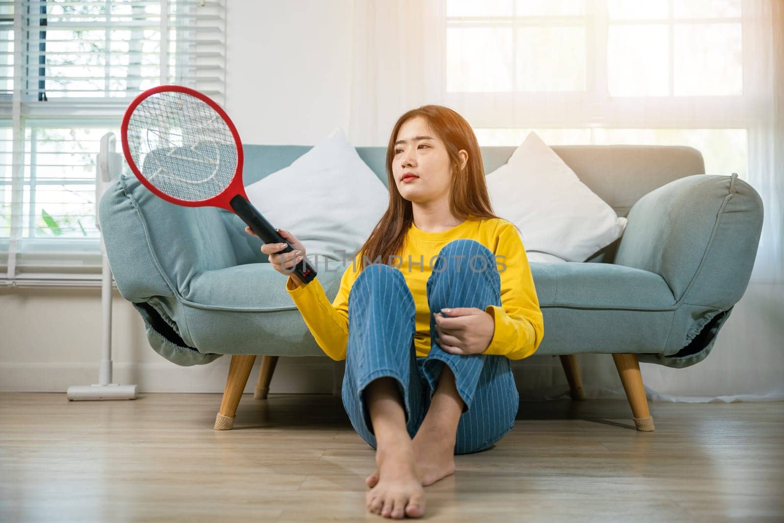 Woman killing mosquitoes hand holding fly swatter like weapon in living room at home, Asian young female sitting floor using mosquito swatter or electric net racket, technology insect killer
