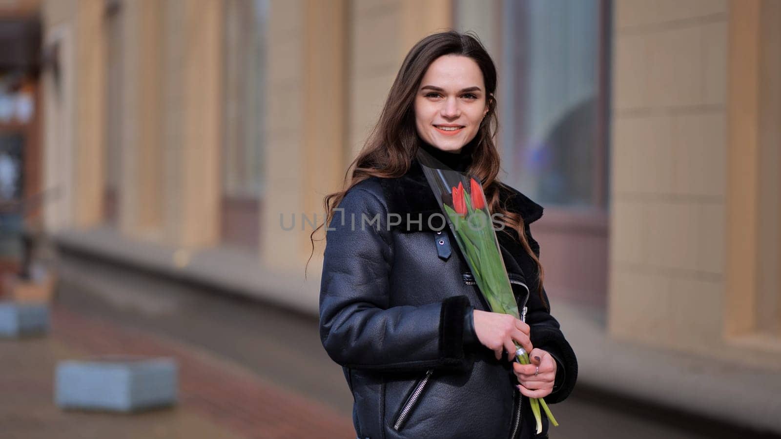 Portrait of a happy brunette girl with a tulip in the background of the city in the cold season. by DovidPro