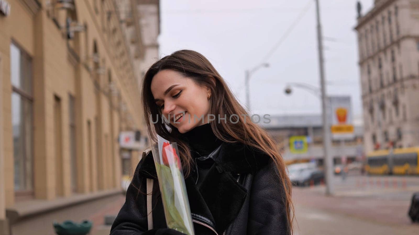 Portrait of a happy brunette girl with a tulip in the background of the city in the cold season. by DovidPro
