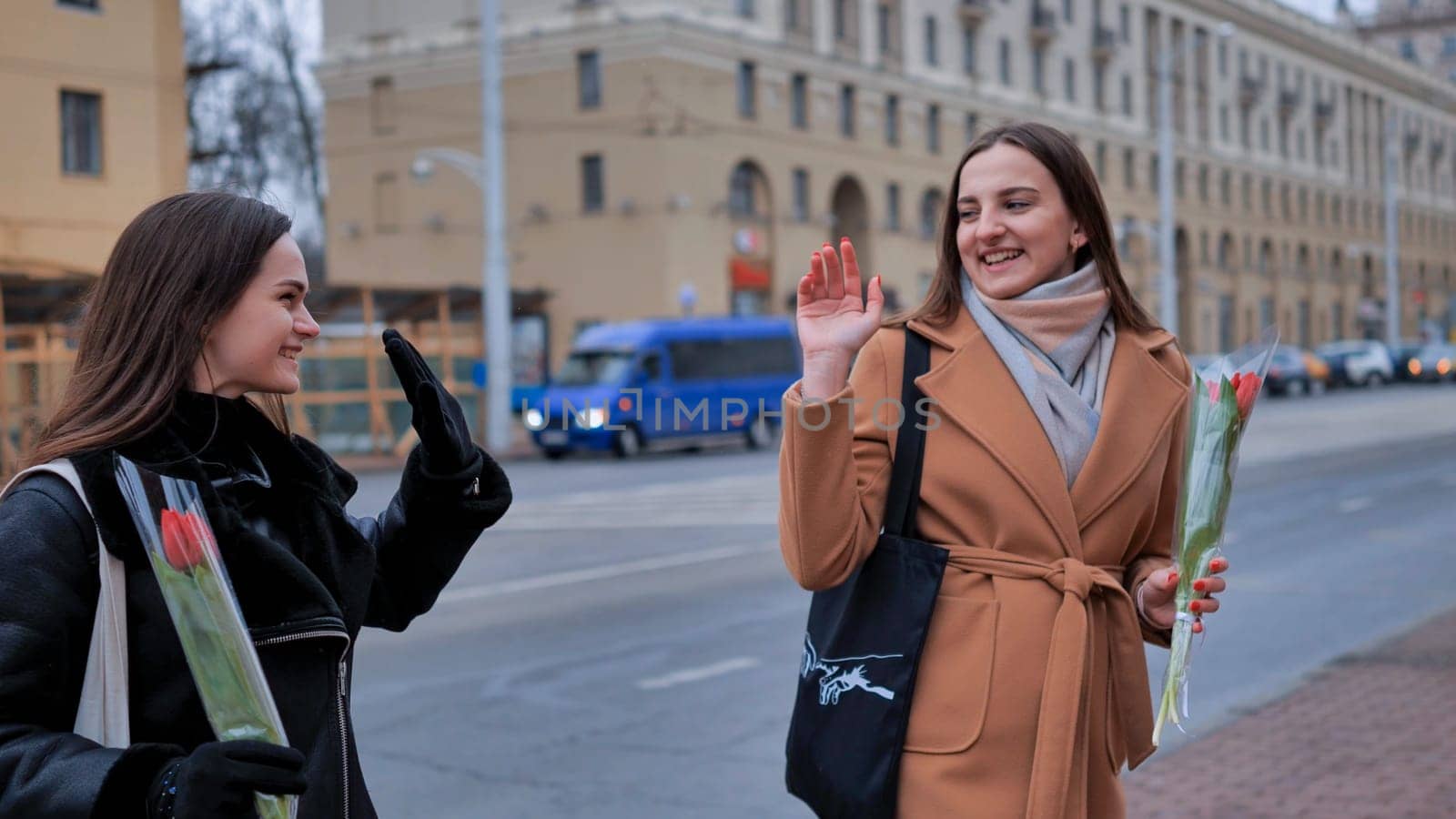 two stylish beautiful young women with flowers in their hands say goodbye on a city street