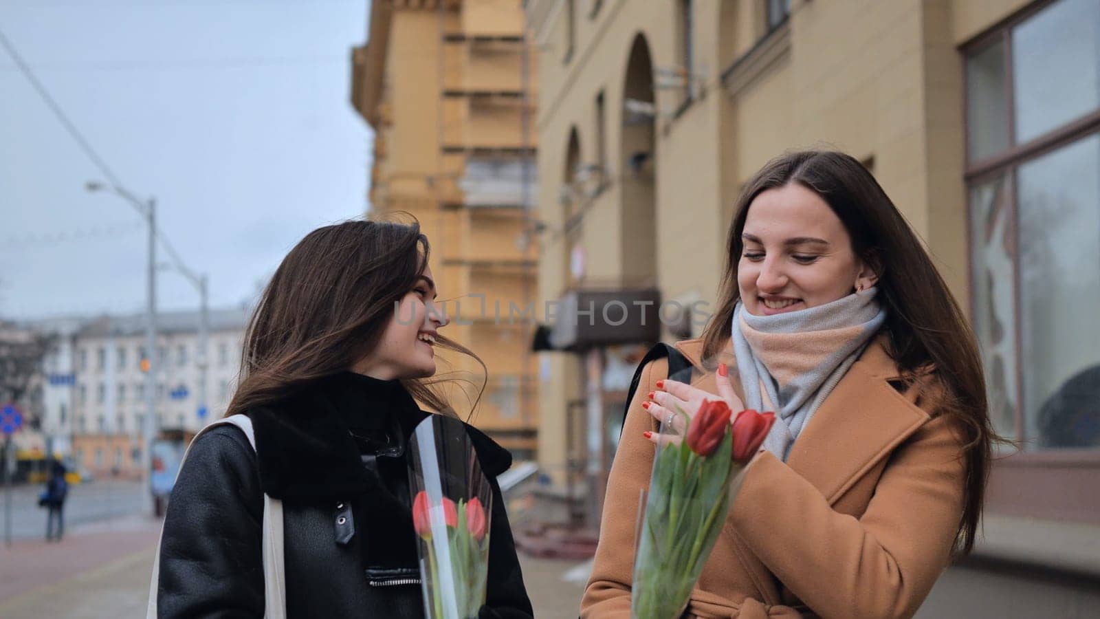 Two young girls, friends with flowers in their hands, walk along a city street on a cloudy spring day.