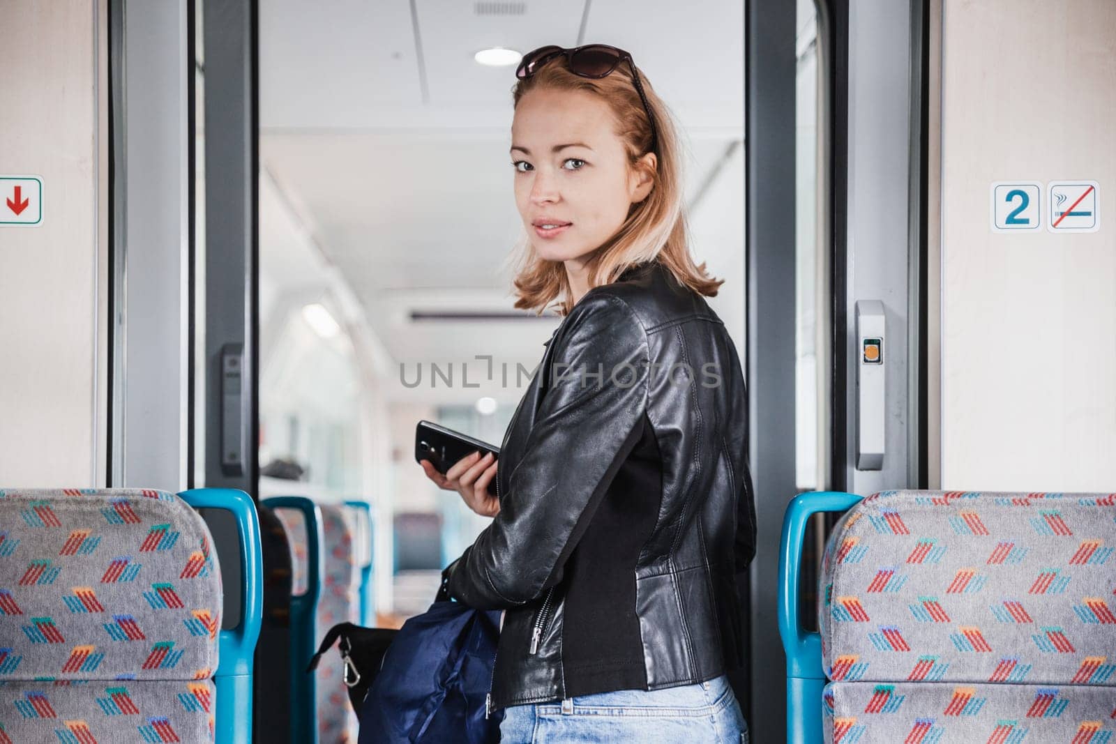 Young blond woman in jeans, shirt and leather jacket holding her smart phone and purse while riding modern speed train arriving to final train station stop. Travel and transportation