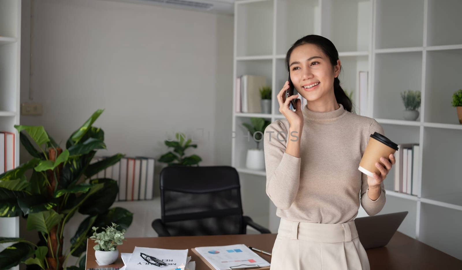 Asian businesswoman is talking on the phone in an online business meeting using a laptop in a modern home office decorated with lush green plants. by wichayada