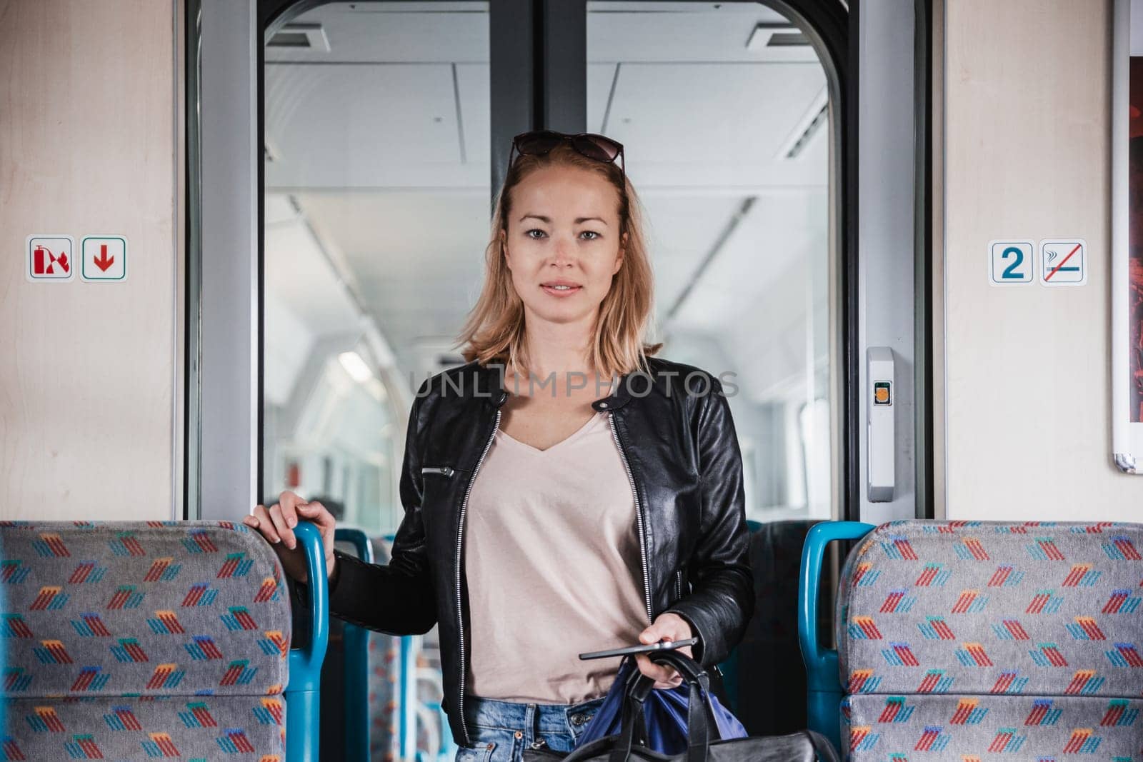 Young blond woman in jeans, shirt and leather jacket holding her smart phone and purse while riding modern speed train arriving to final train station stop. Travel and transportation. by kasto