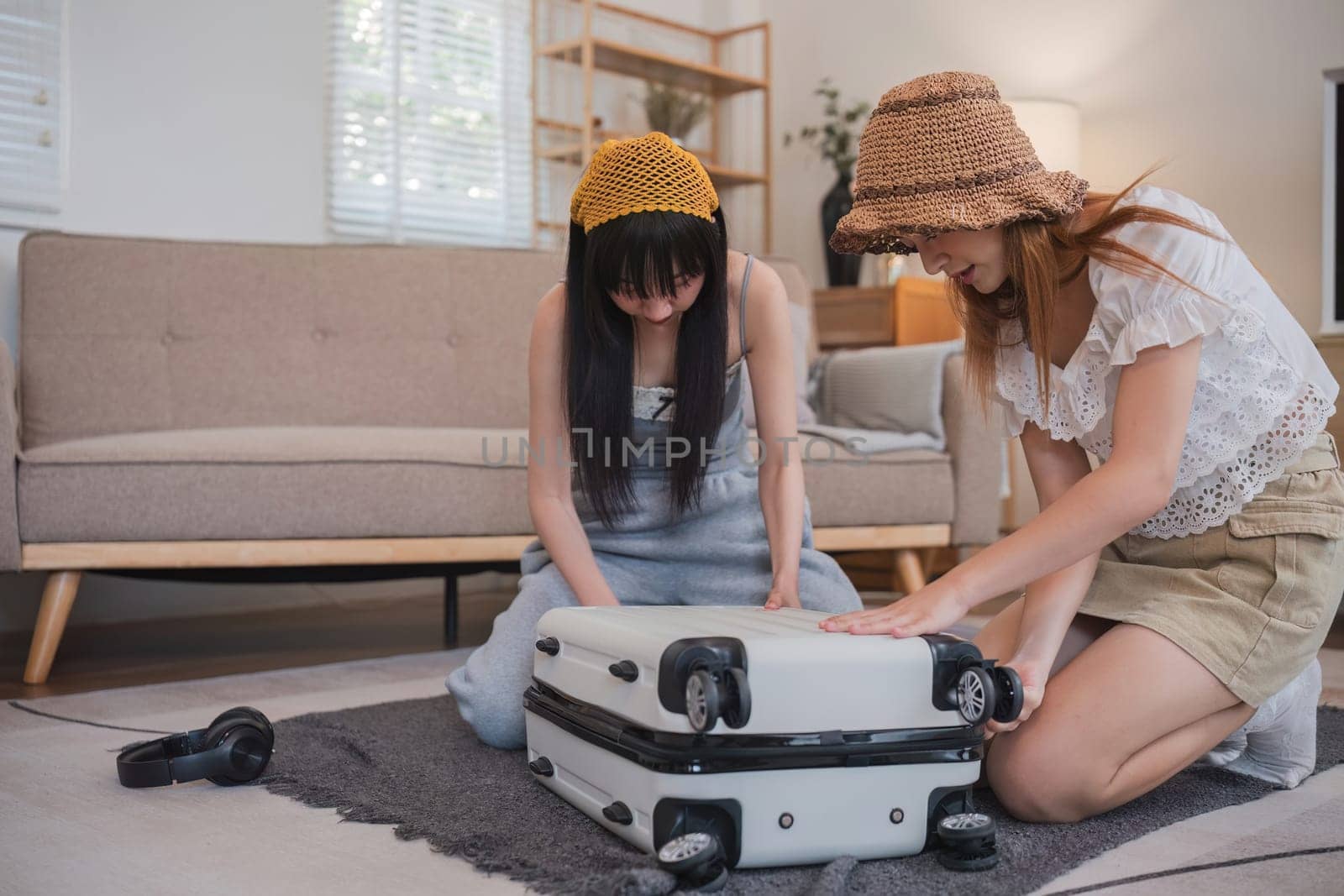 Two women are looking at a suitcase on the floor. One of them is wearing a yellow hat