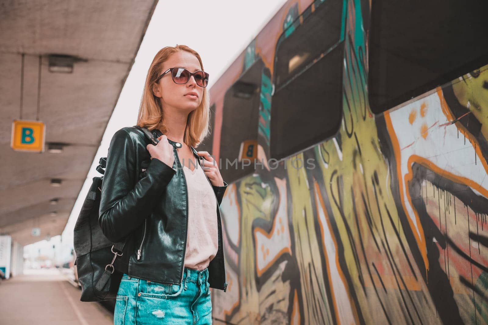 Young blond woman in jeans, shirt and leather jacket wearing bag and sunglass, embarking modern speed train on train station platform. Travel and transportation. by kasto