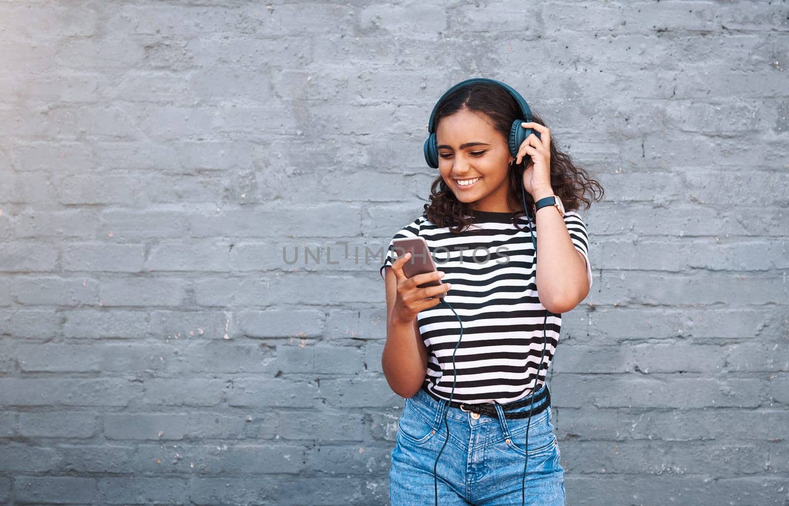 Girl, headphones and cellphone with wall background for music listening, subscription or podcast. Female person, smile and urban city with technology for entertainment with playlist, app or mockup by YuriArcurs