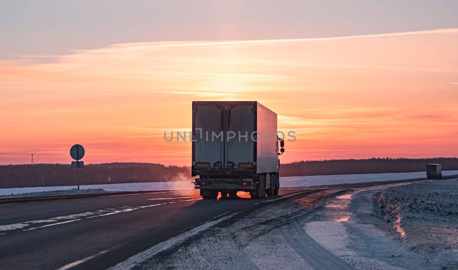 A semi truck cruises down a wintry highway as the sun sets on the horizon, casting a warm glow over the icy road and surrounding landscape.