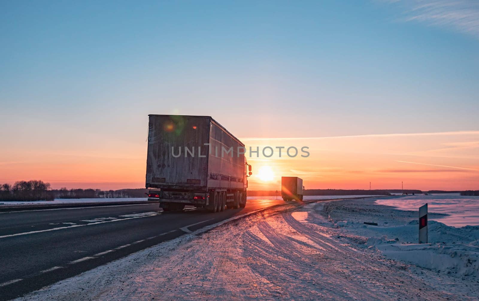 A semi truck cruises down a wintry highway as the sun sets on the horizon, casting a warm glow over the icy road and surrounding landscape.