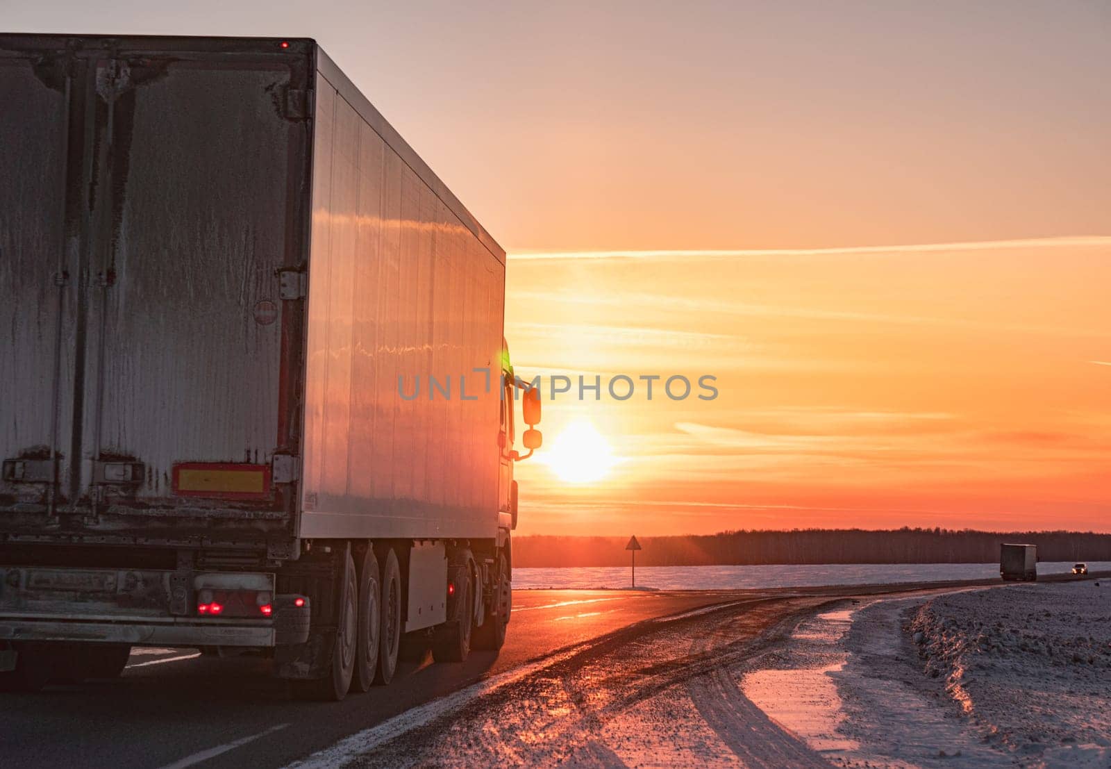 A semi truck cruises down a wintry highway as the sun sets on the horizon, casting a warm glow over the icy road and surrounding landscape.