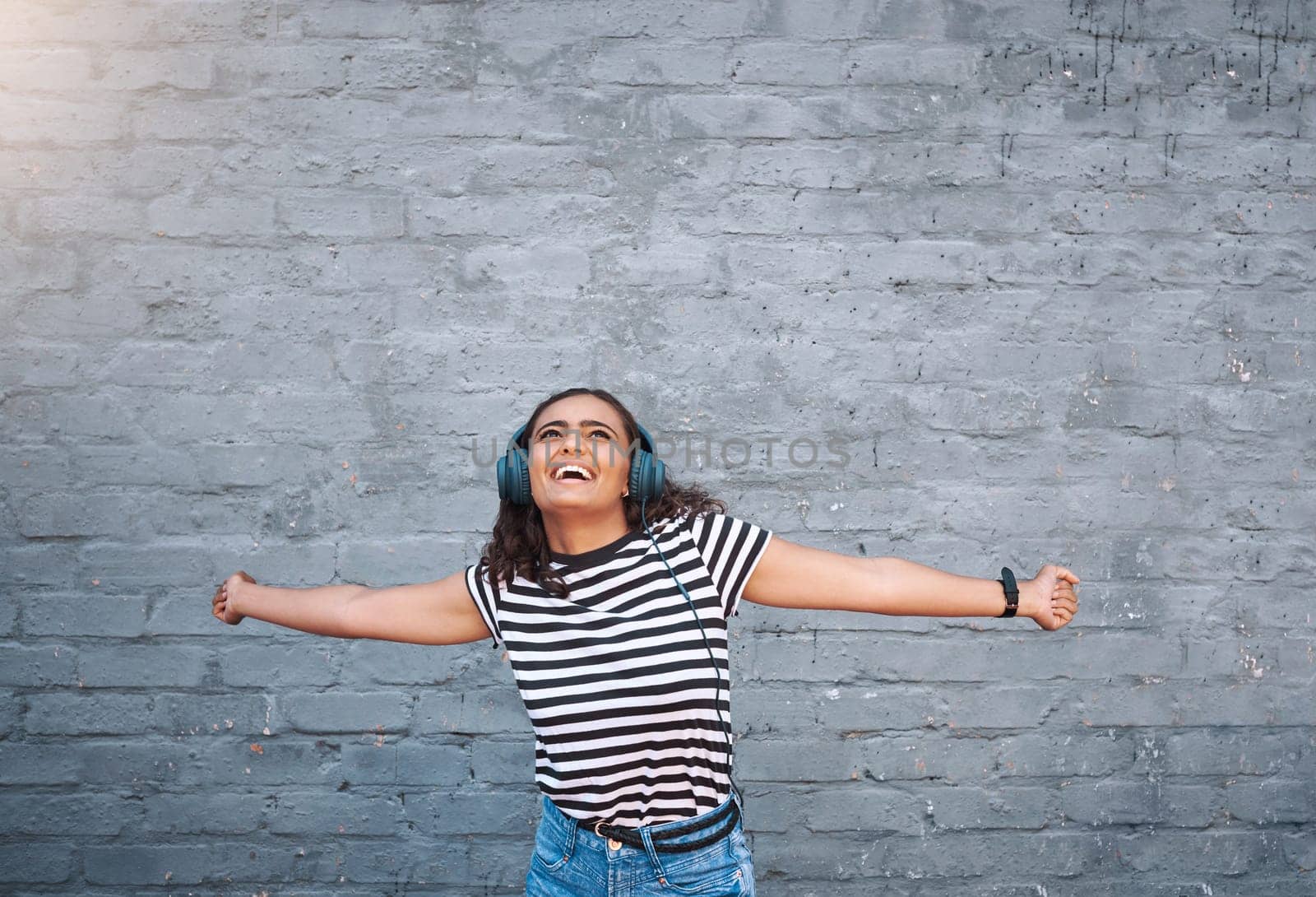 Happy, excited and girl with headphones, streaming music and smile on grey wall with mindset, joyful and cheerful. Person, outdoor and headset for radio, listening to audio and sound with freedom by YuriArcurs