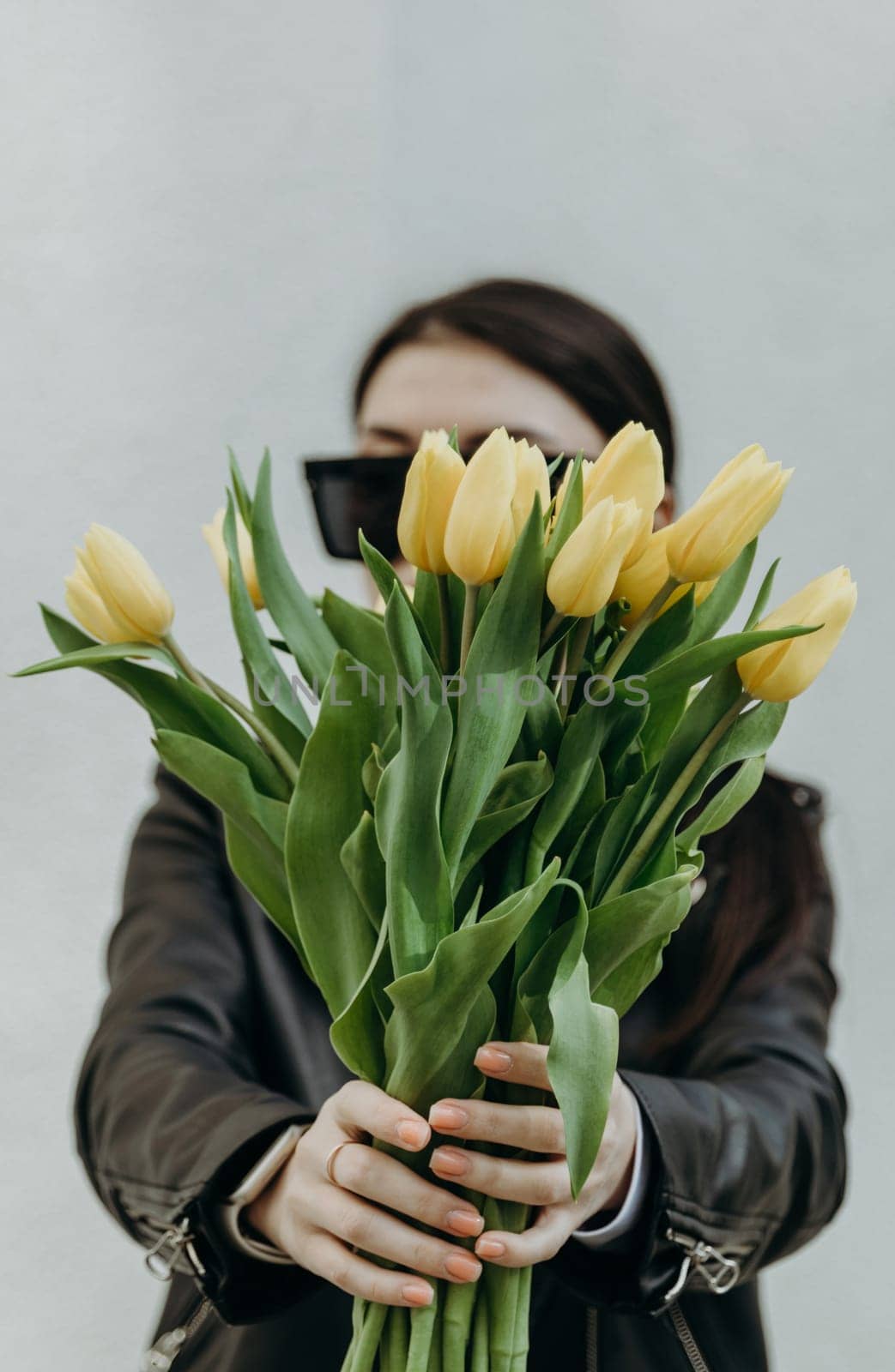 Portrait of one young beautiful Caucasian girl in sunglasses, leather jacket stretching her hands forward with a bouquet of yellow tulips, covering her face, standing near a white wall on a spring day and looking down, side view close-up.
