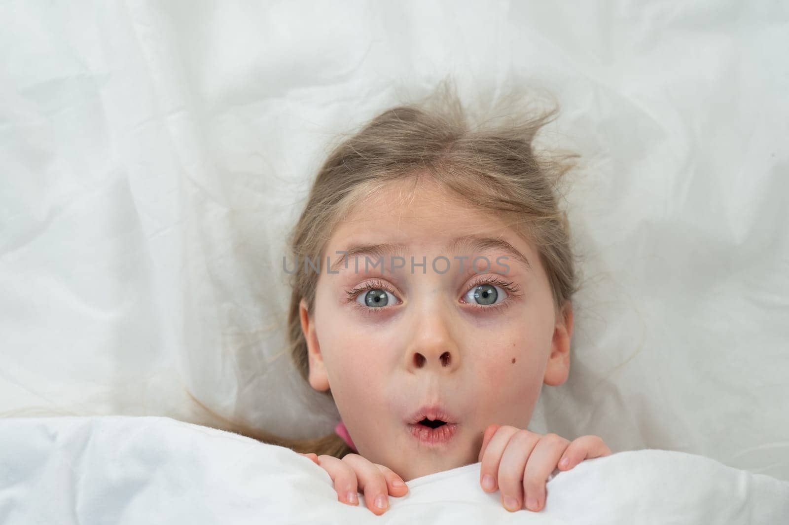 Top view of a grimacing little girl lying in bed