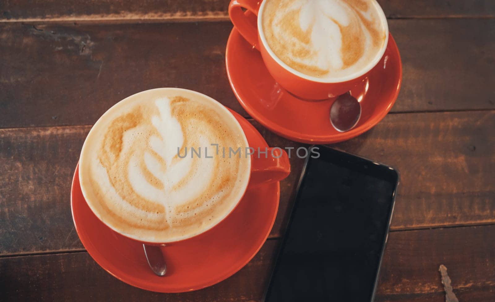Top view of two red cups of coffee with latte art on wooden desktop with the smartphone. Table in cafe.