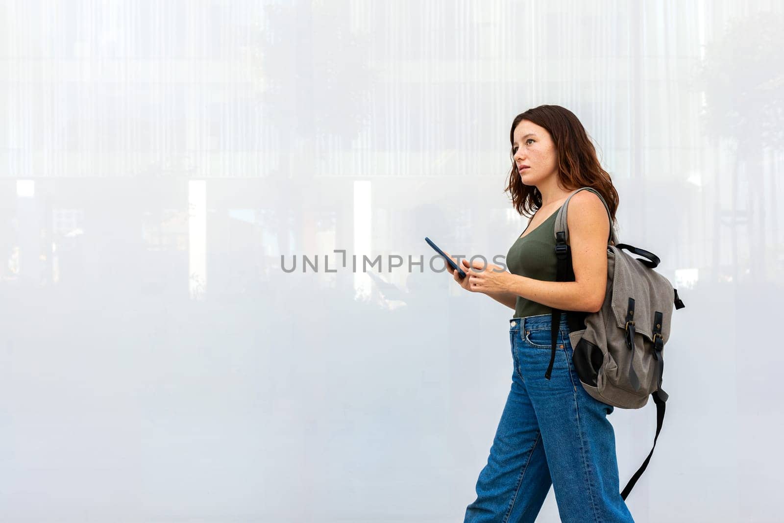 Female university student with backpack walking to class in college campus. Young redhead woman in city street using mobile phone. Copy space. Education and technology concepts.