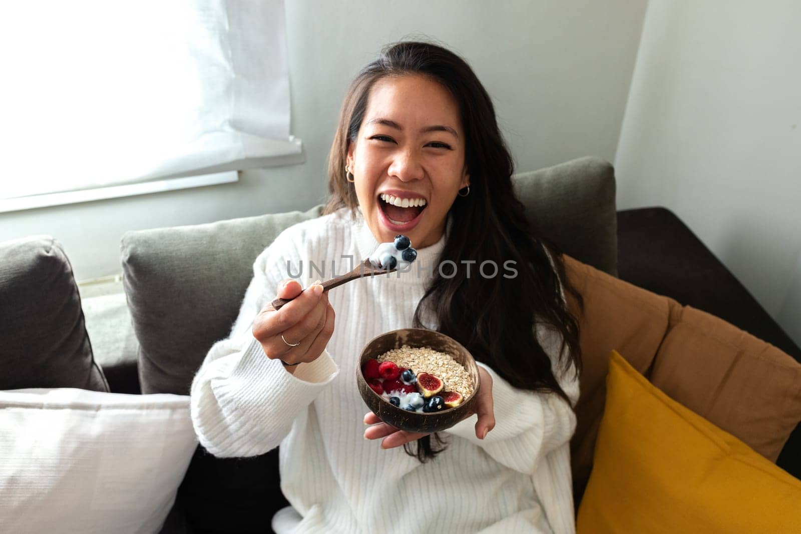 Smiling young happy Asian woman eating healthy breakfast bowl of yogurt looking at camera. Healthy lifestyle concept.