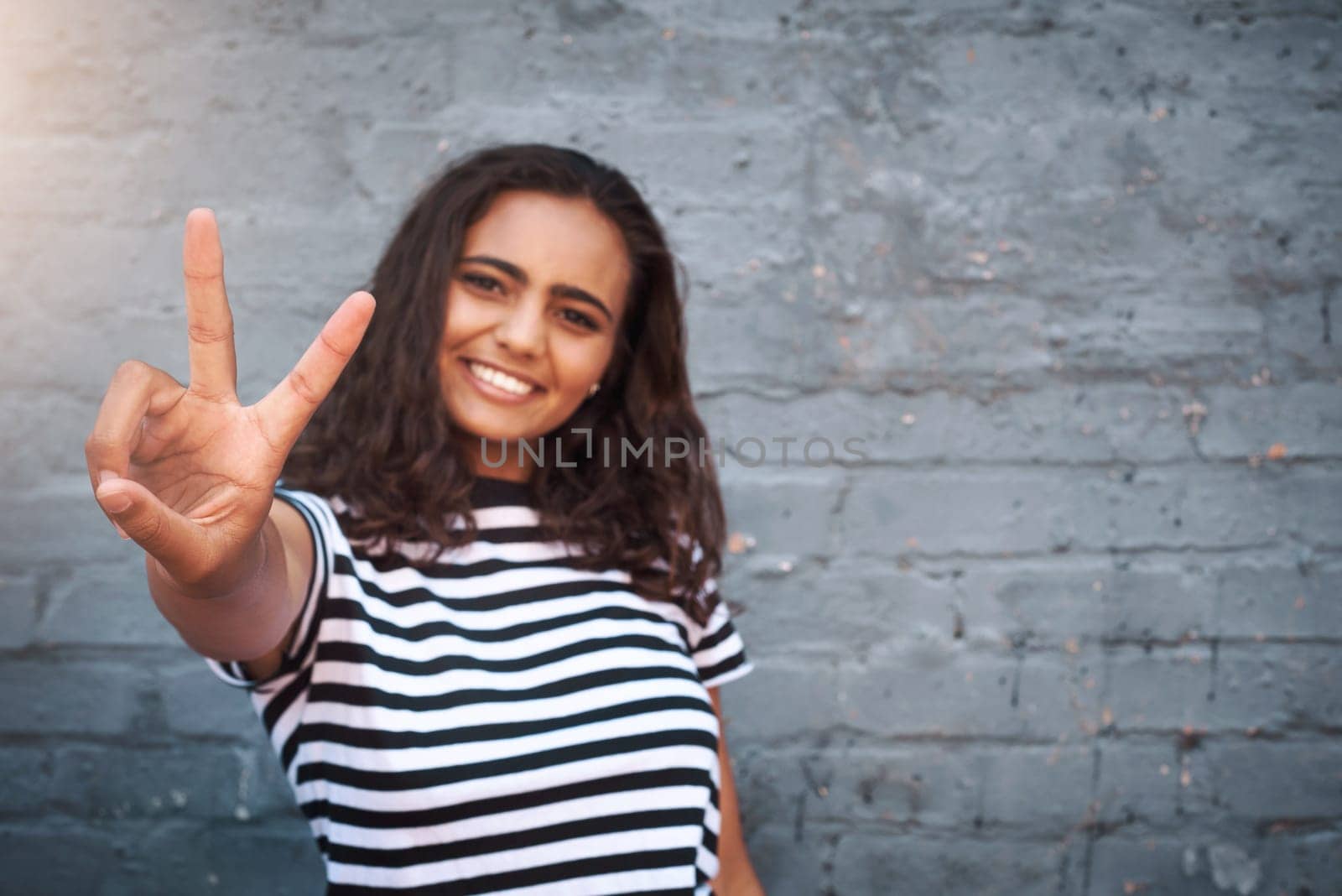 Happy girl, portrait and peace sign with wall background for emoji, positivity or fun attitude. Young female person or friendly hipster with smile, gesture or fashion for urban style on mockup space by YuriArcurs