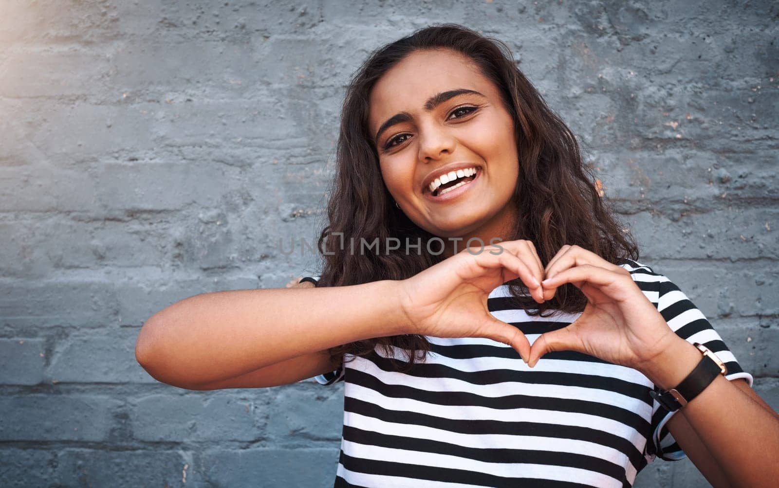 Brick, wall and heart sign for girl, smile and hands for gesture, outdoor and break for weekend. Proud, excited and student on holiday, morning and happiness on face, model and city of New York.