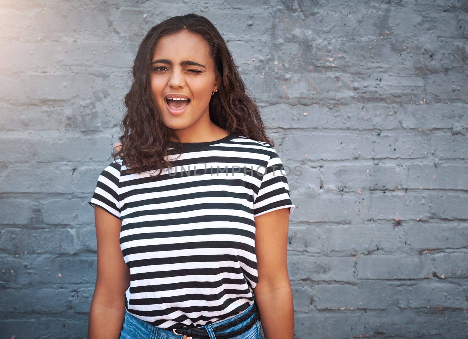 Brick, wall and girl with wink, happy and student in university with scholarship, goal and education. Outdoor, campus and college of Germany, smile and academy for learning and portrait of person.