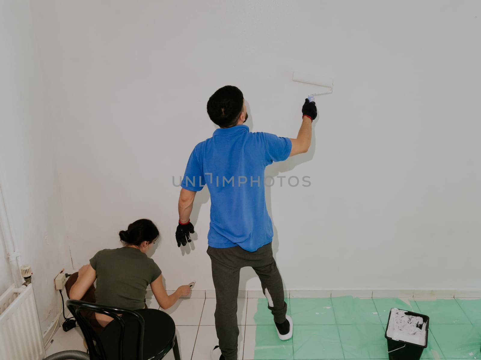 A young handsome dark-haired Caucasian man in a blue T-shirt and a girl stand on a tiled floor with a green oilcloth from the back and paint a wall with white paint with a roller, close-up side view with selective focus.