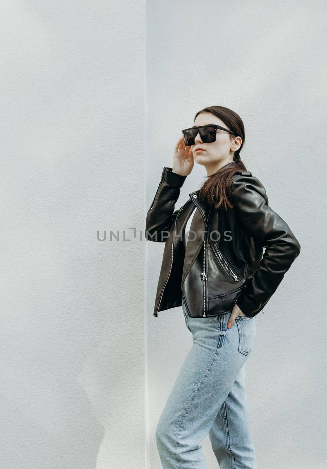 Portrait of one young beautiful Caucasian girl in sunglasses, leather jacket and blue jeans standing sideways and posing to the right near a white wall on a spring day with copy space on the left, close-up bottom view.