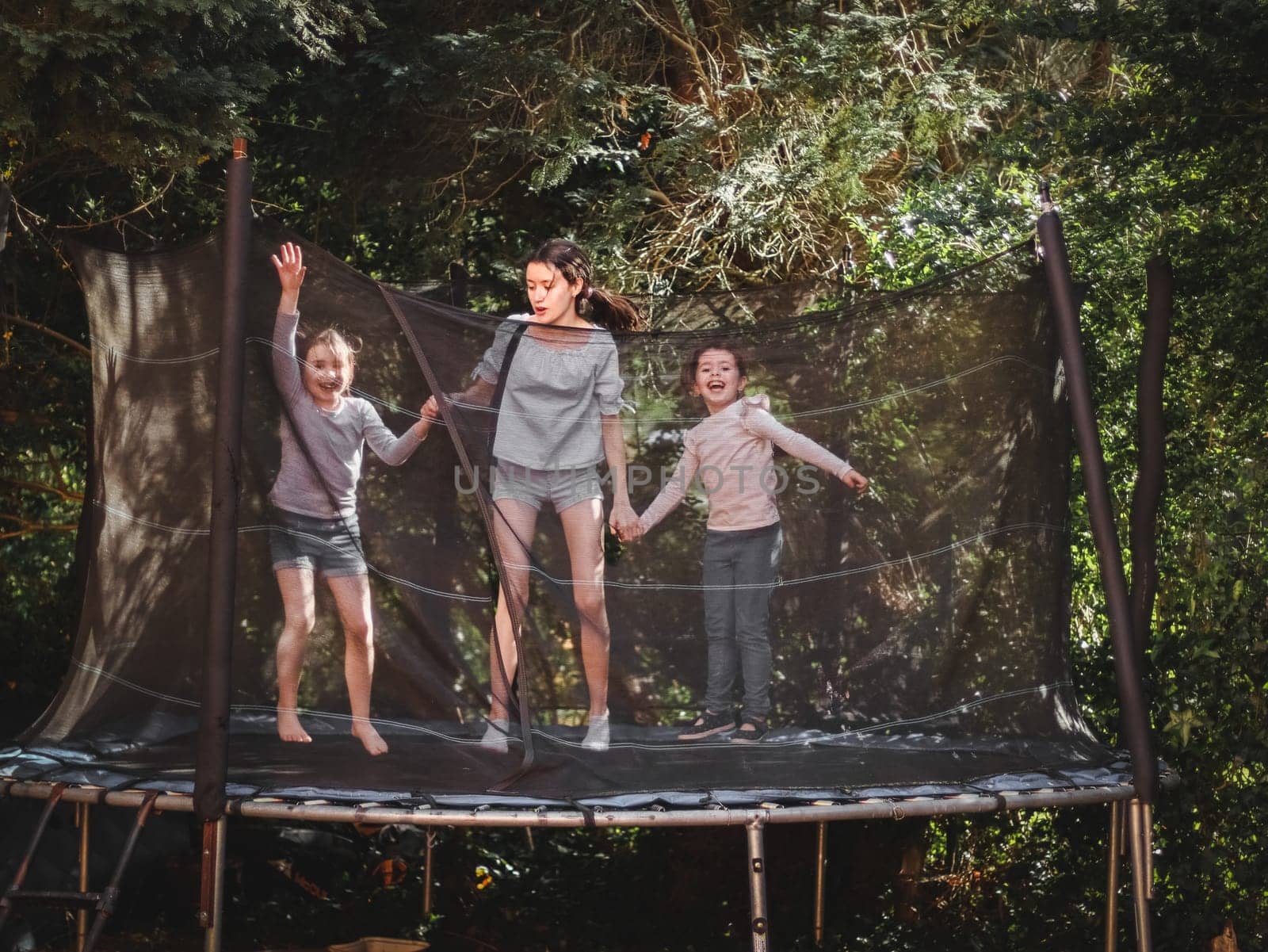 Three beautiful caucasian sister girls in jeans, shorts and a sleeved t-shirt jump on a trampoline, frozen in happy with smiles on their faces in flight in the garden of a house among trees, close-up side view. Concept at home, home sports, happy childhood, children entertainment.